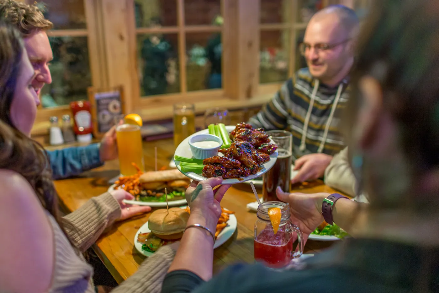 Group of people sitting at a wooden table sharing plates of chicken wings, celery, a sandwich, and drinks in a cozy restaurant setting.