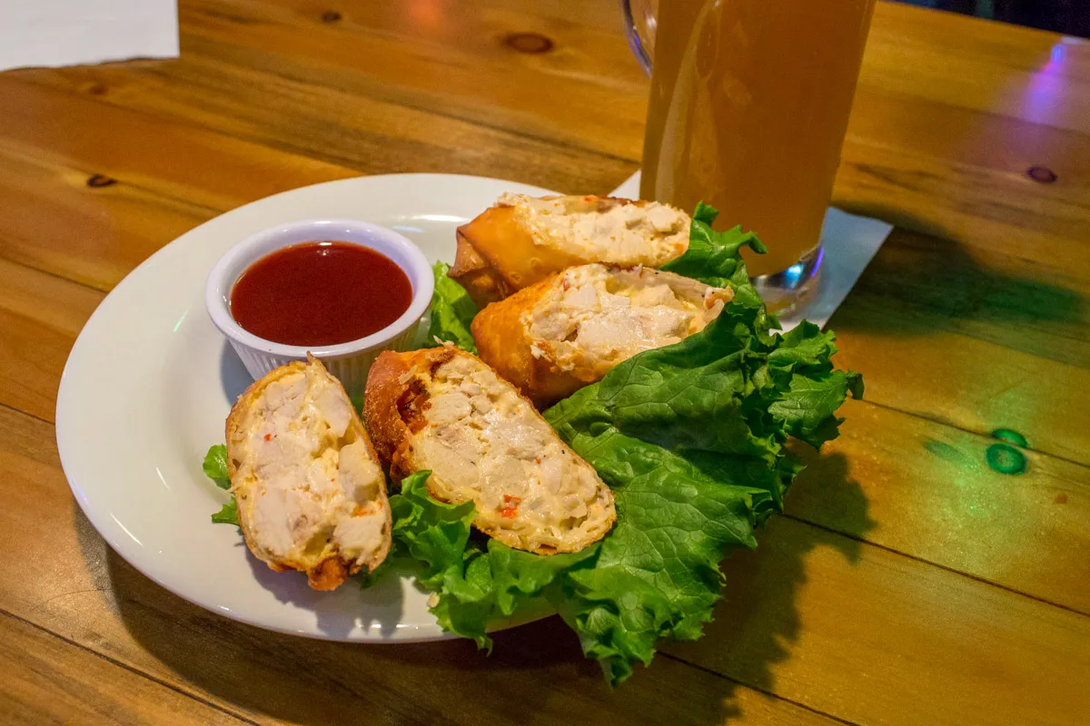 Plate with fried crab rangoons on green leaf lettuce and a small cup of red dipping sauce, set on a wooden table with a glass of iced tea in the background.