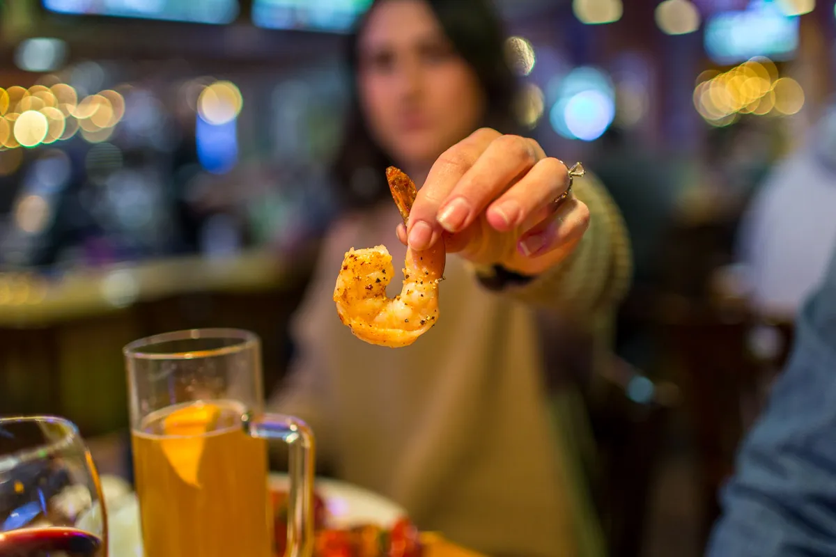 Person holding a seasoned cooked shrimp toward the camera in a dimly lit restaurant or bar setting.