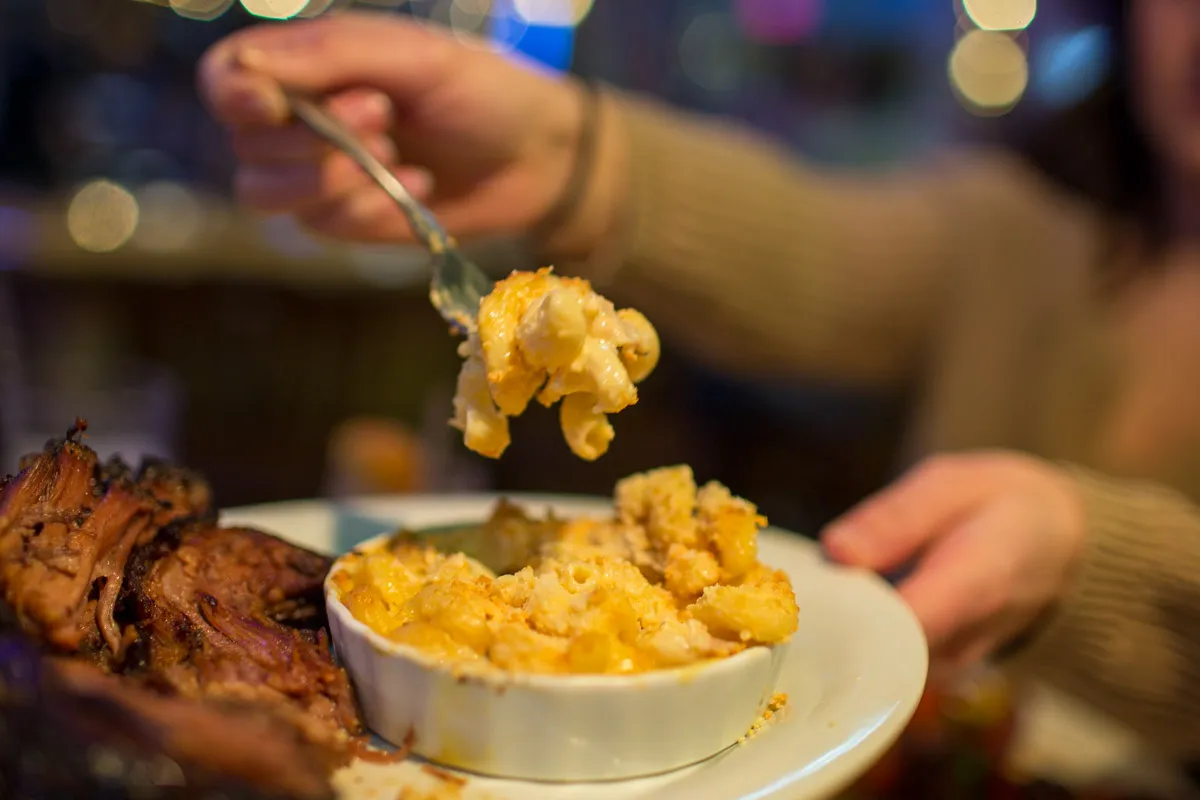 Person holding a forkful of cheesy baked macaroni and cheese above a plate with more macaroni in a white dish and barbecue ribs.