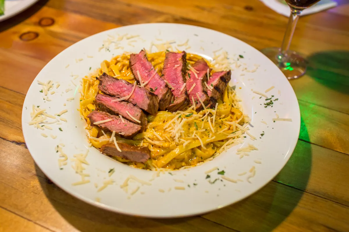 Plate of fettuccine pasta with creamy sauce topped with slices of medium-rare steak and sprinkled with grated cheese and herbs.