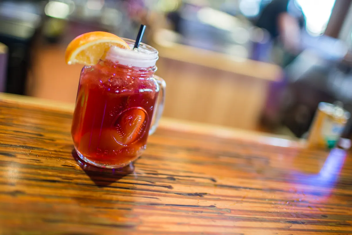 Glass jar filled with a red iced drink garnished with a slice of orange and a black straw on a wooden table.