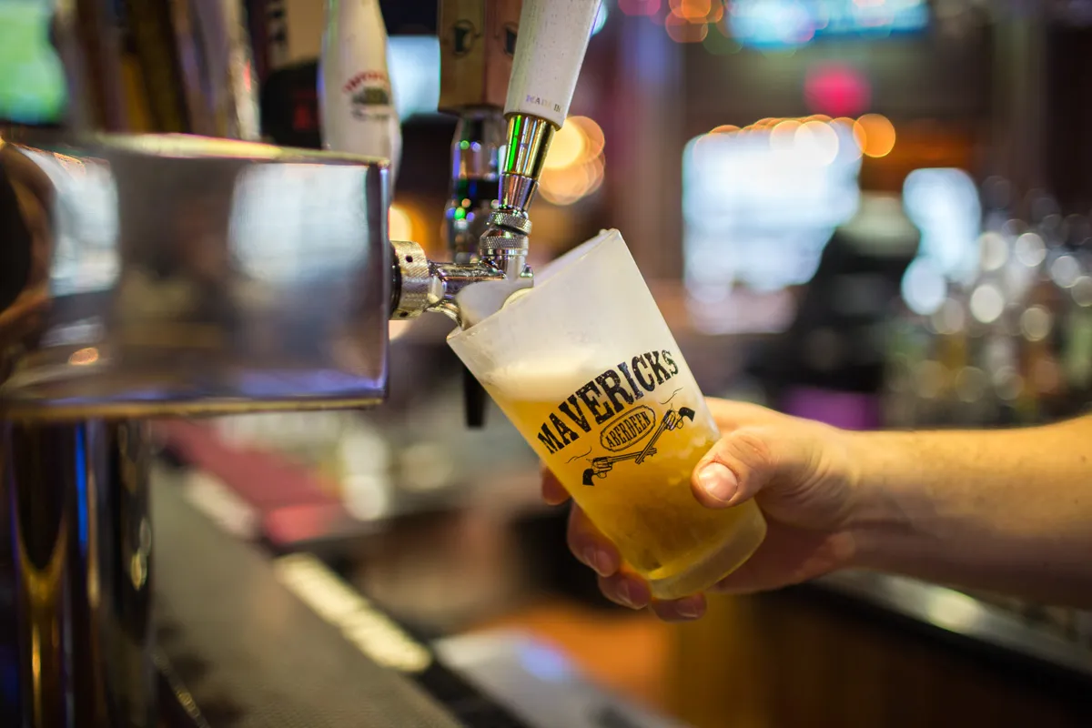 Hand holding a frosted glass with 'Mavericks Aberdeen' logo, filling with draft beer from a tap in a bar.