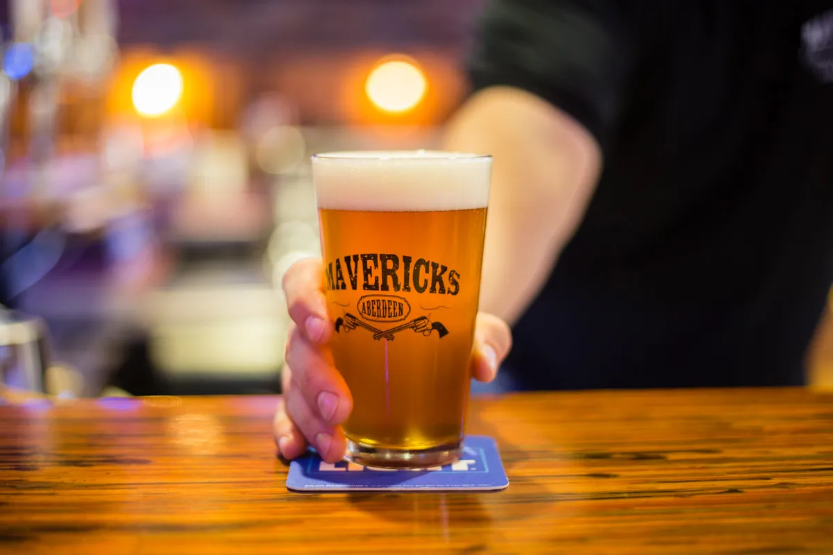 Hand holding a glass of amber beer with foam on top labeled 'Mavericks Aberdeen' on a wooden bar counter.