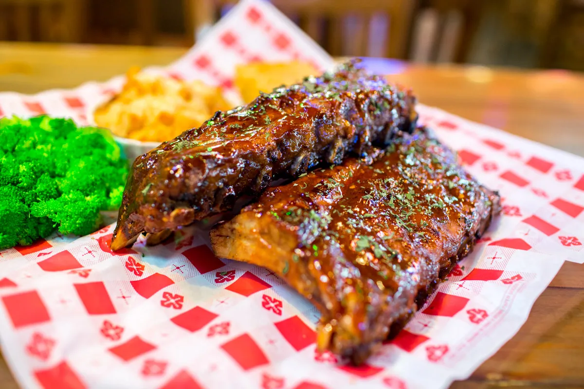 Barbecue pork ribs glazed with sauce served with steamed broccoli and macaroni and cheese on checkered paper.
