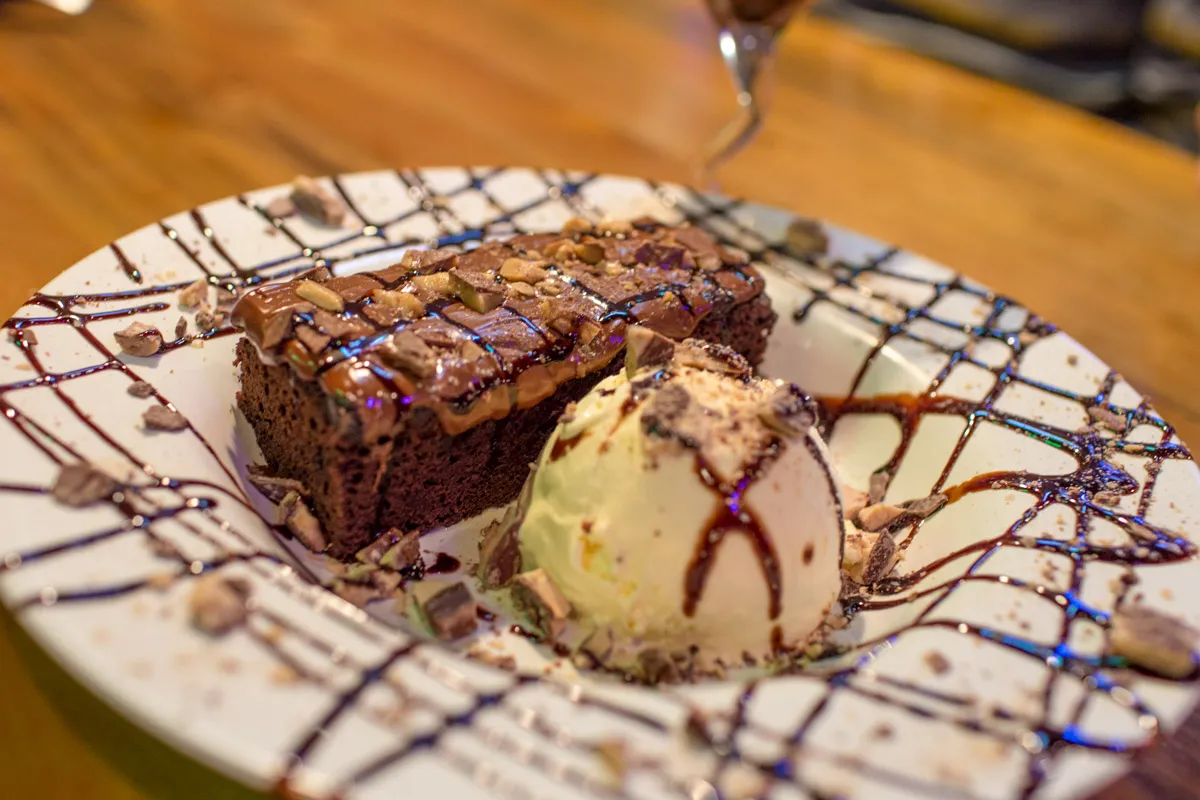 Plate with a slice of nut-topped chocolate cake and a scoop of vanilla ice cream drizzled with chocolate syrup.