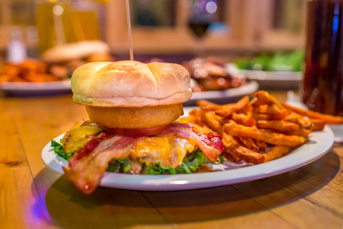 Bacon cheeseburger with lettuce, tomato, onion ring, and a side of sweet potato fries on a white plate.