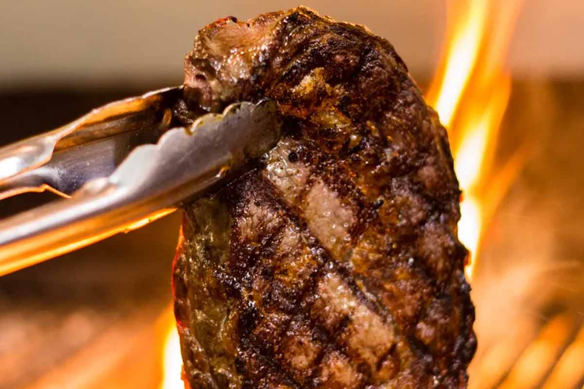 Close-up of a grilled steak held by metal tongs with flames in the background.