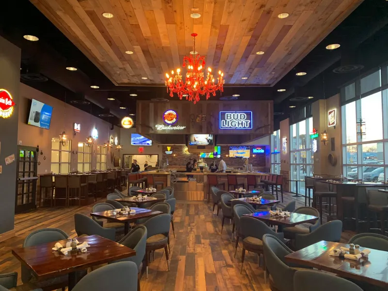 Spacious bar and restaurant interior with wooden floors and ceiling, tables with chairs, a red chandelier, and illuminated beer brand signs including Budweiser and Bud Light.