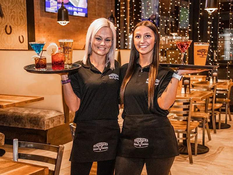 Two female waitresses smiling and holding trays with drinks in a cozy restaurant interior.