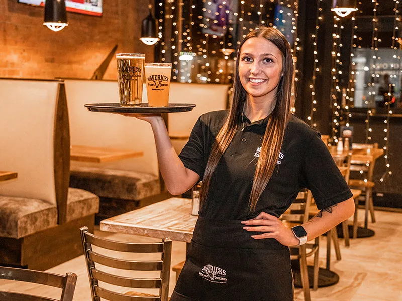 Smiling waitress holding a tray with two drinks inside a warmly lit restaurant with string lights and wooden furniture.