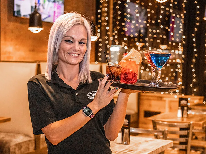 Smiling waitress with light purple hair holding a tray with three colorful cocktails in a warmly lit restaurant.