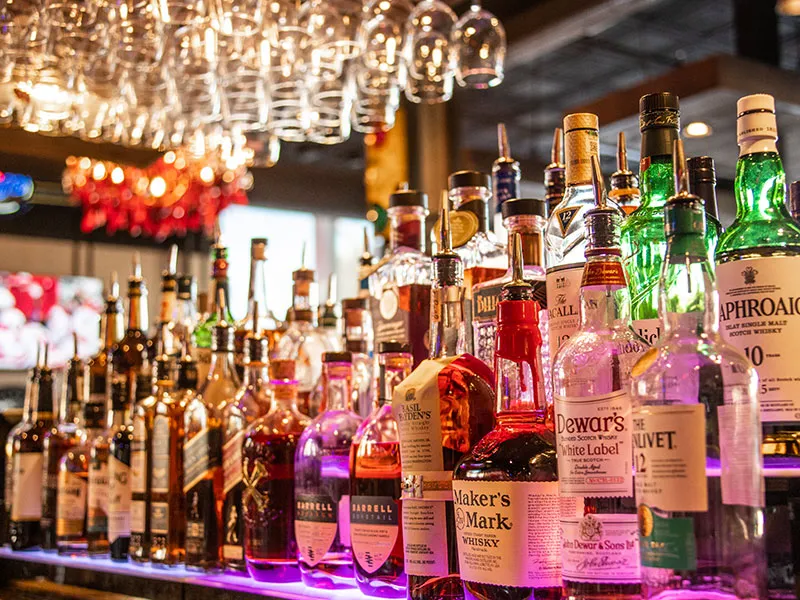 Bar shelf with various whiskey bottles and glasses hanging upside down above, illuminated by purple lighting.