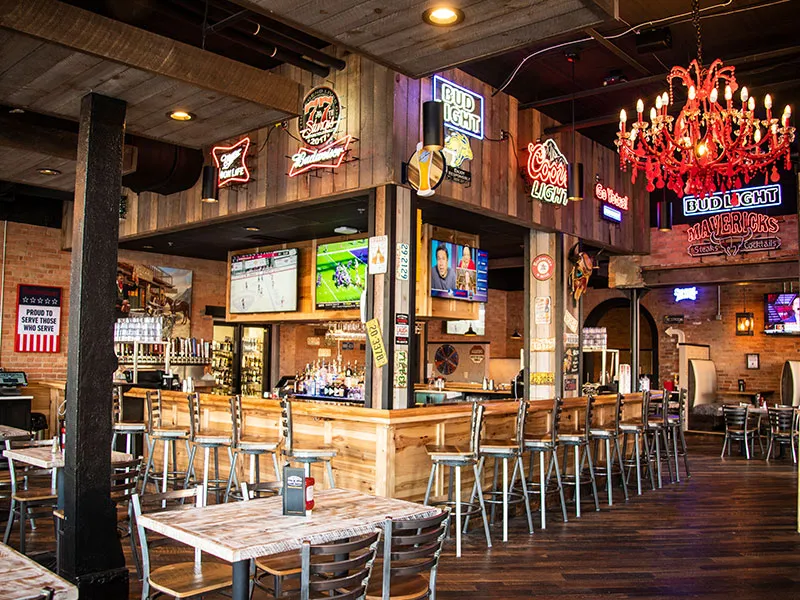 Interior of a lively bar with a wooden counter surrounded by high stools, multiple TV screens, neon beer signs, and a red chandelier.