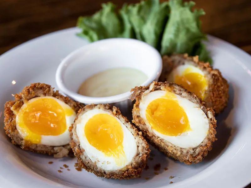 Plate with four halved Scotch eggs featuring soft yolks, a leafy lettuce garnish, and a small bowl of dipping sauce.