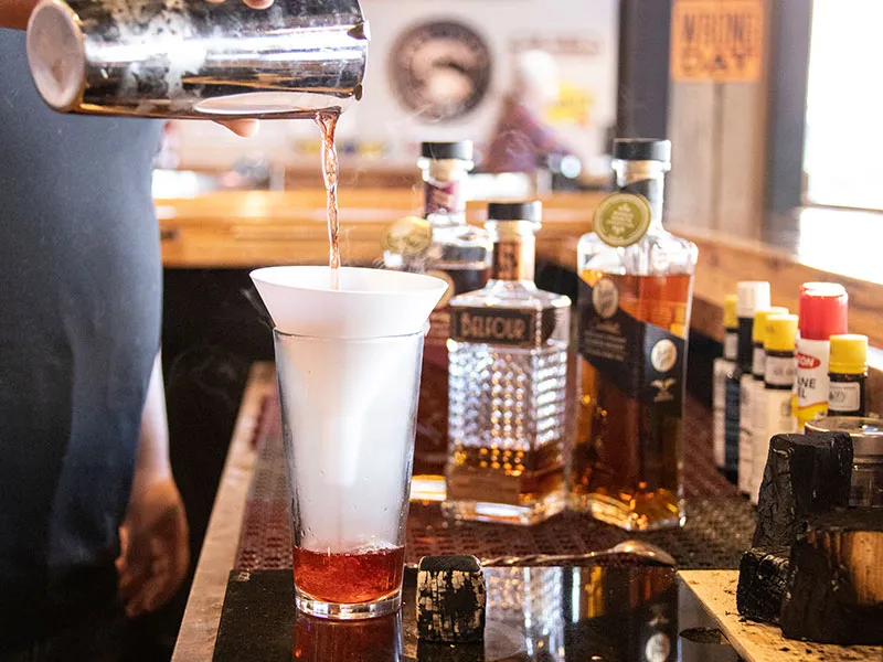 Bartender pouring a drink through a white funnel into a tall glass with a red liquid base on a bar counter with bottles and drink ingredients in the background.