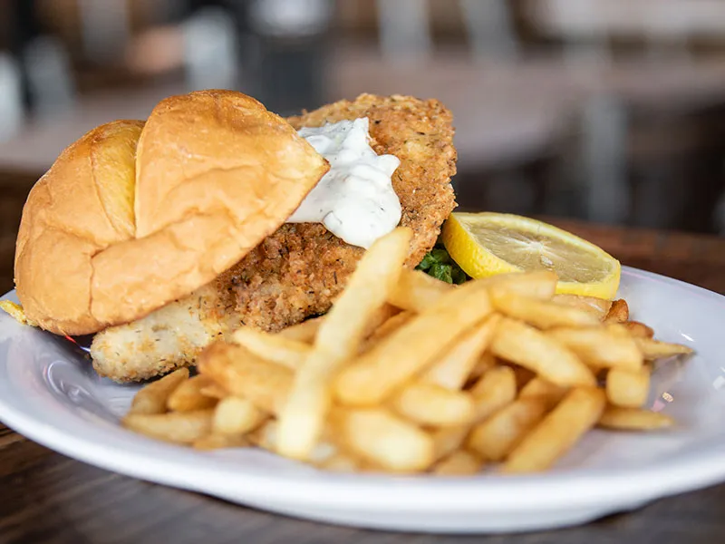Plate with fish sandwich topped with tartar sauce, a lemon wedge, and French fries on a wooden table.