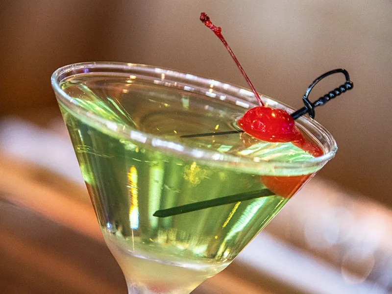 Close-up of a green cocktail in a martini glass garnished with a red cherry on a black cocktail pick.