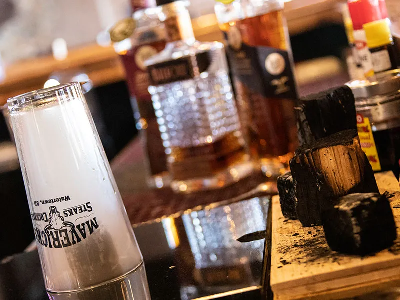Close-up of a smoking glass with vapor inside, next to charred wood pieces and blurred whiskey bottles on a wooden surface.