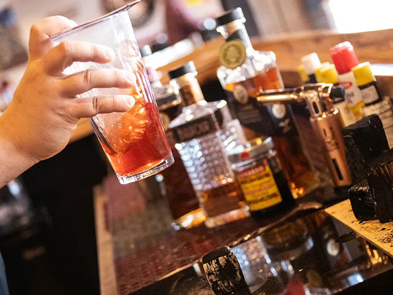Hand holding a glass with a red cocktail and ice in front of a bar counter lined with various liquor bottles and a lighter.