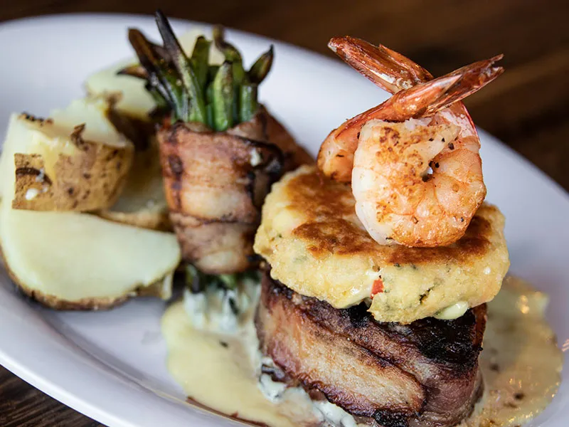 Plate with steak topped with a crab cake and grilled shrimp, served with bacon-wrapped green beans and potato wedges.