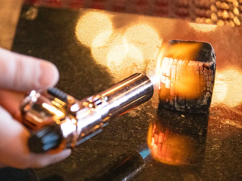 Hand holding a small torch lighter igniting a glowing charred charcoal cube on a reflective surface.