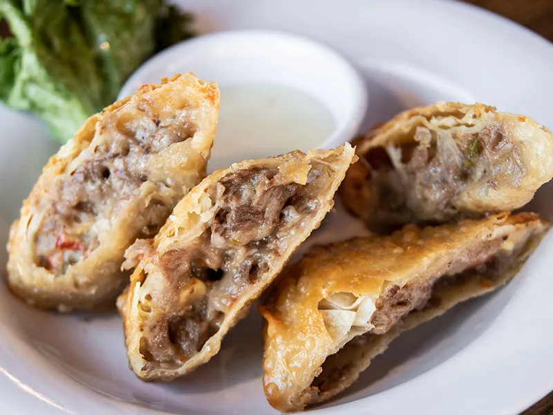 Plate with four fried egg rolls cut in half revealing a meaty filling and a small bowl of white dipping sauce.