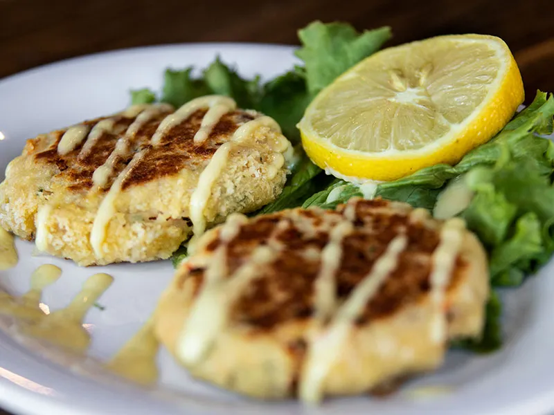 Two golden-brown crab cakes drizzled with sauce on a bed of leafy greens with a lemon wedge on a white plate.