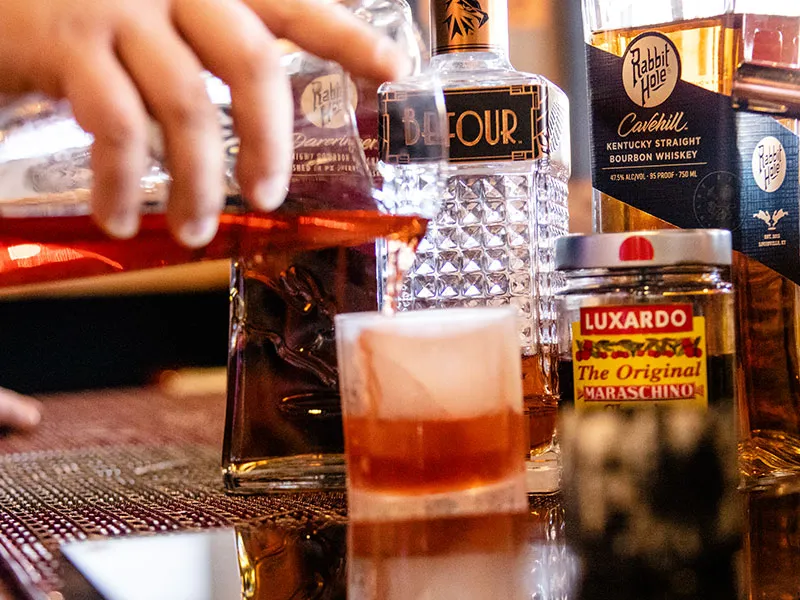 Hand pouring a reddish-brown cocktail into a glass with a large ice cube, surrounded by bottles of BAFour, Rabbit Hole Cavehill bourbon, and Luxardo maraschino cherries.