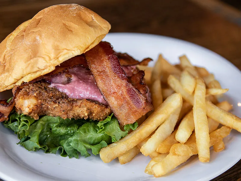 Fried chicken sandwich with bacon and lettuce on a bun served with French fries on a white plate.