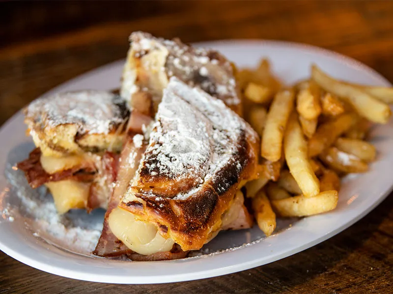 Plate with toasted ham and melted cheese sandwich dusted with powdered sugar and a side of french fries on a wooden table.