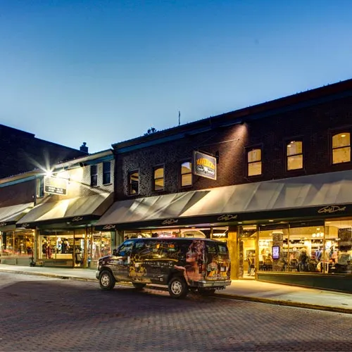 Evening street view of a brick building with warmly lit shops and a decorated van parked in front.