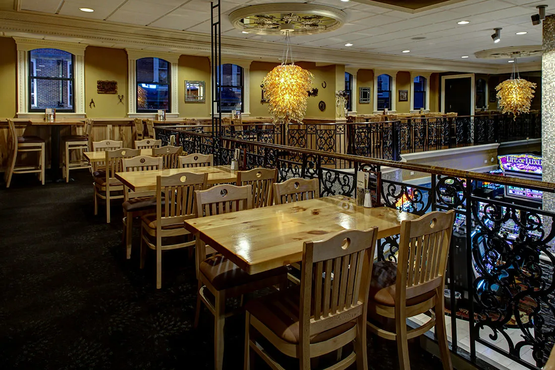 Interior of a dimly lit restaurant with wooden tables and chairs, decorative iron railing, and hanging chandeliers.
