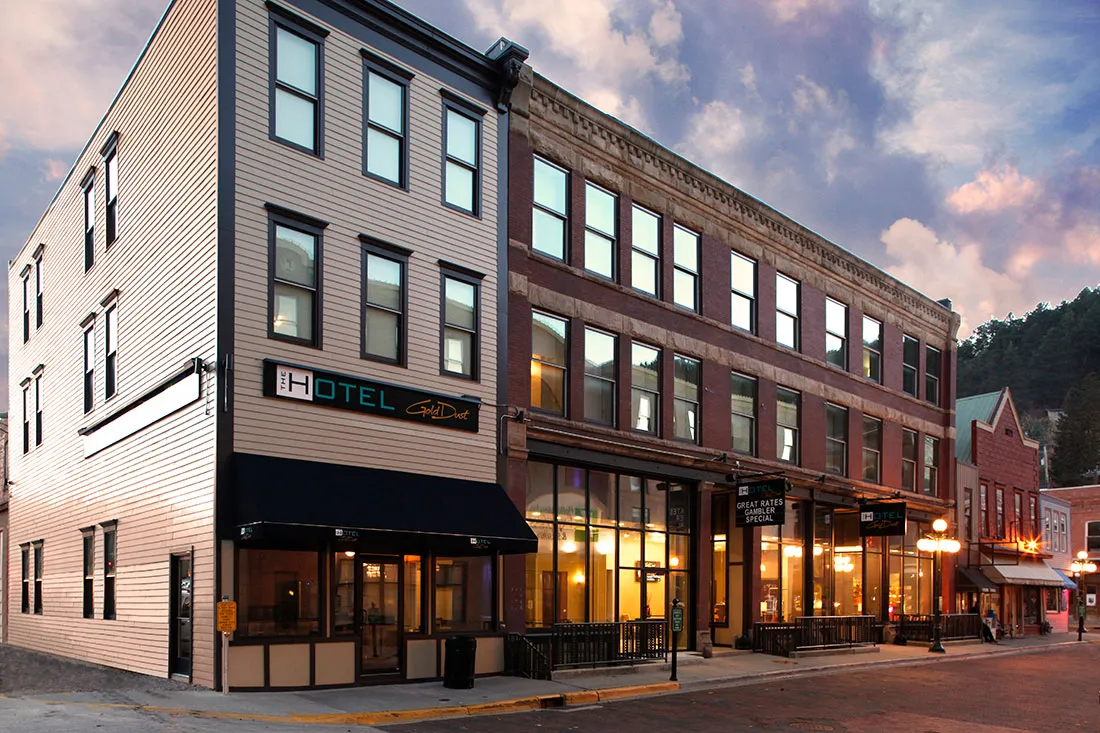 Exterior view of a three-story hotel building named 'The Hotel Gold Dust' on a street corner at dusk with illuminated windows and a purple-pink cloudy sky.