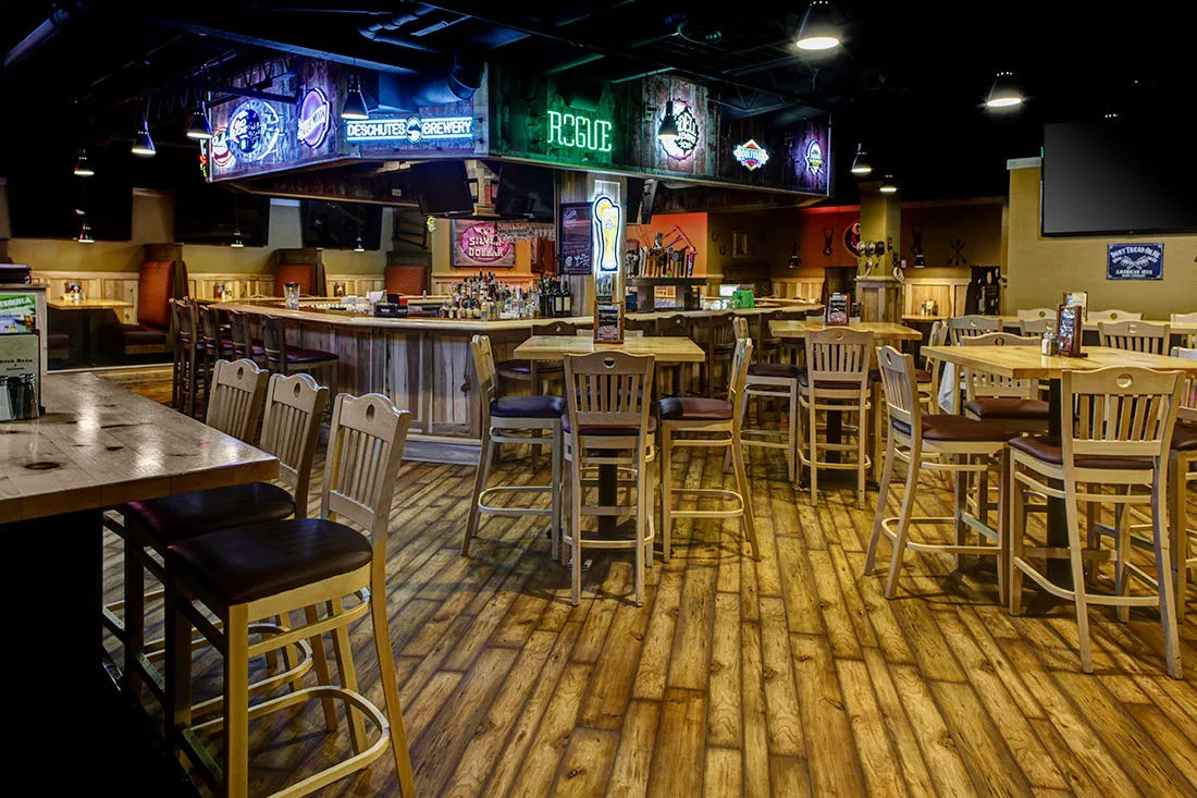 Empty rustic bar with wooden floors, high chairs, tables, and illuminated beer brand signs overhead.