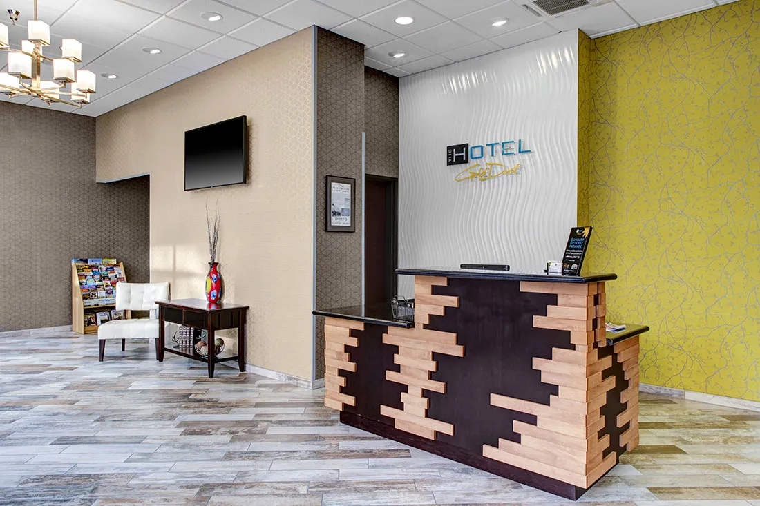 Hotel reception area with a modern wooden front desk, patterned walls, a seating area with a white chair and table, and a wall-mounted TV.