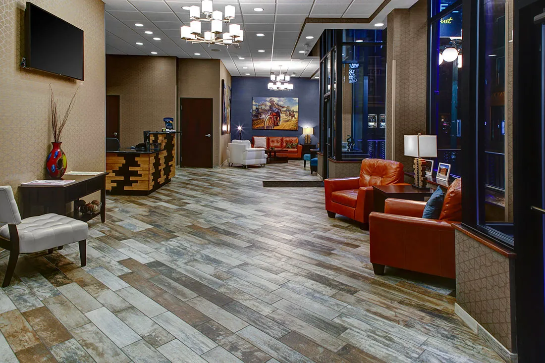 Modern hotel lobby with wooden patterned floor, tan walls, orange leather chairs, white armchairs, reception desk, and large windows showing a night street view.