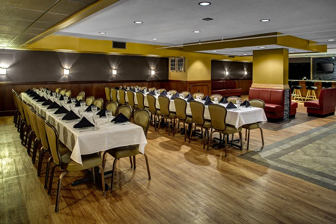 Banquet room with two long tables set with white tablecloths, black napkins, glassware, and beige chairs, adjacent to a seating area with red booths and bar stools.