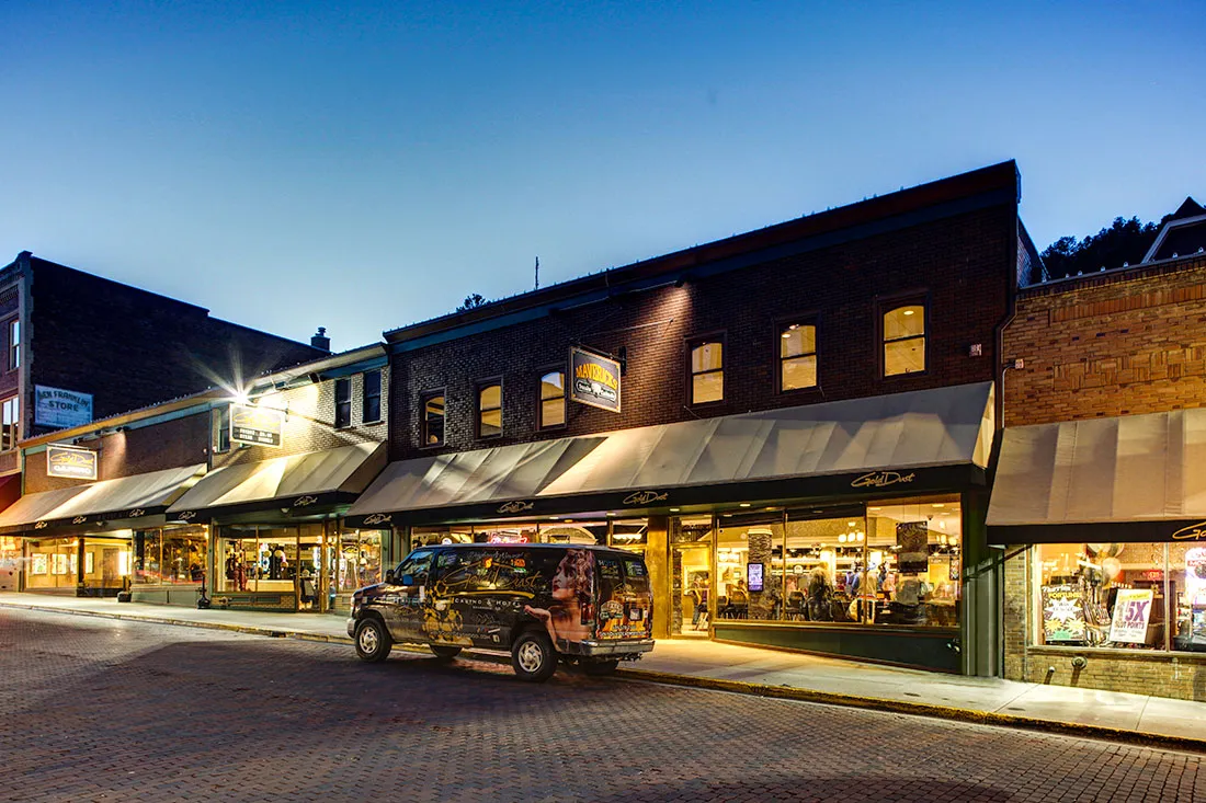 Evening street view of a row of shops with large windows and awnings, including a Gold Dust store with a parked promotional van in front.