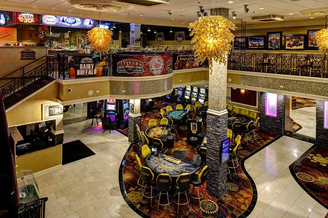 Interior of a casino with gaming tables, slot machines, yellow chairs, decorative lighting, and a mezzanine level featuring bar signs and framed pictures.