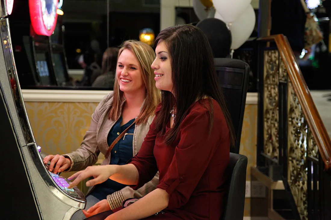 Two women sitting side by side playing a slot machine at a casino.