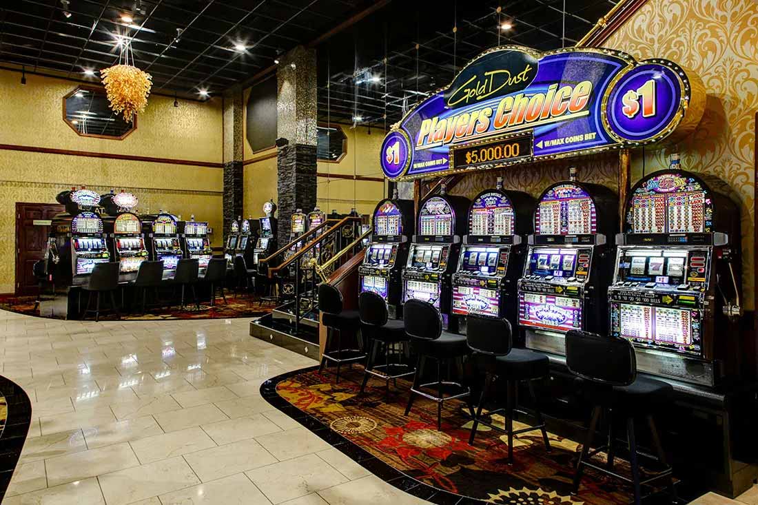 Row of slot machines with chairs in a casino featuring a large illuminated Gold Dust Players Choice $1 sign.