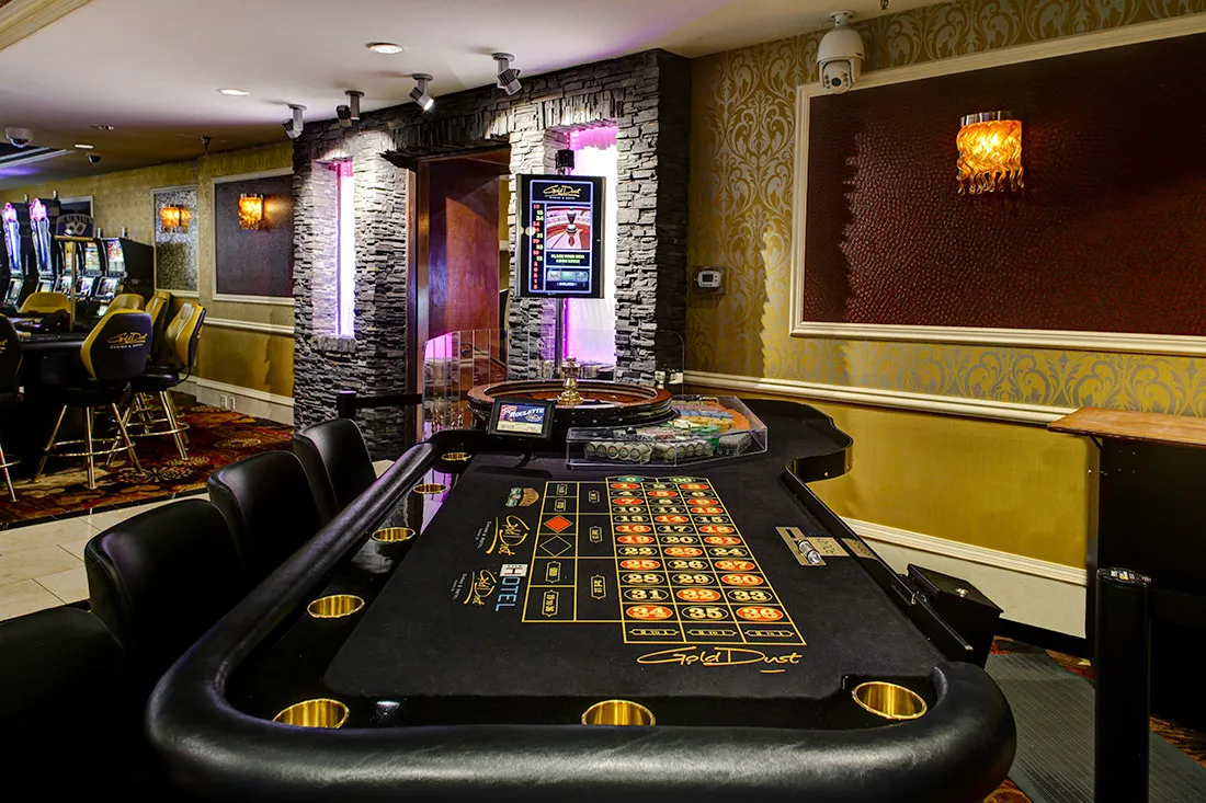 Empty roulette table with black leather chairs and illuminated casino slot machines in a warmly lit gaming room.
