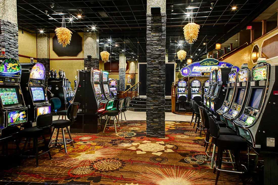 Interior of a casino with rows of slot machines on both sides and decorative hanging lights above.