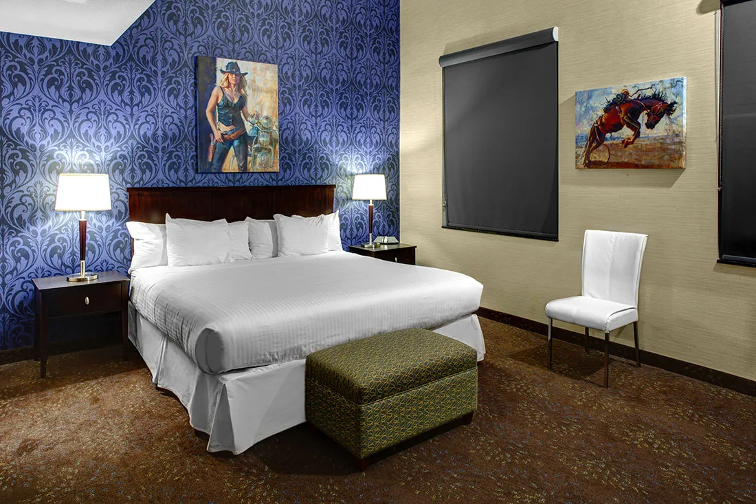 Hotel room with a king-size bed, white linens, blue patterned accent wall, two bedside lamps, an ottoman, and western-themed artwork.