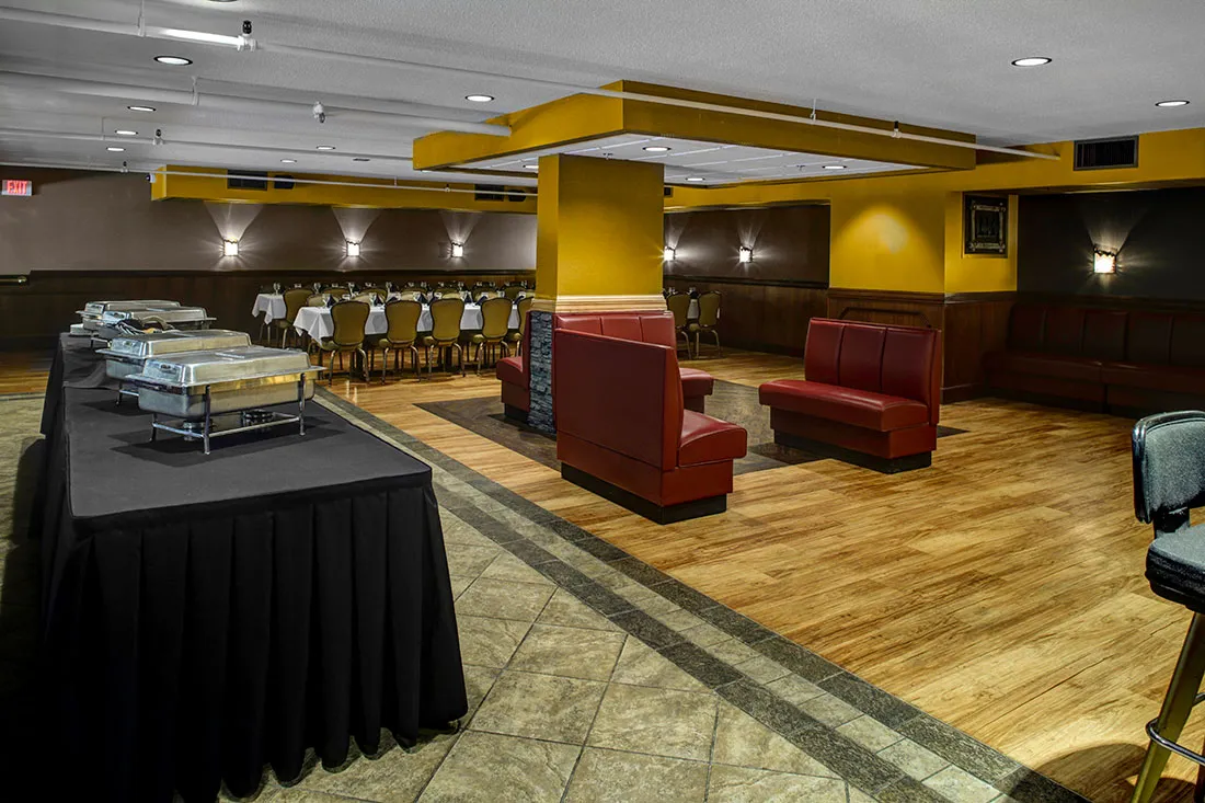 Indoor dining area with a buffet table covered in black cloth and silver chafing dishes, red leather booth seating, wooden and tiled flooring, and a long table set with chairs and white tablecloths in the background.