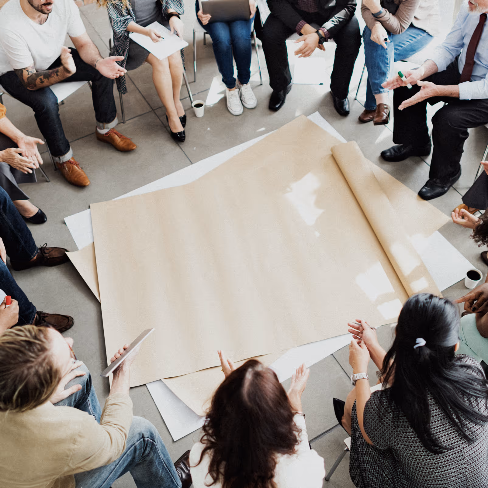 Gruppe von Menschen sitzt im Kreis um einen auf dem Boden ausgebreiteten leeren Papierbogen bei einem Workshop.