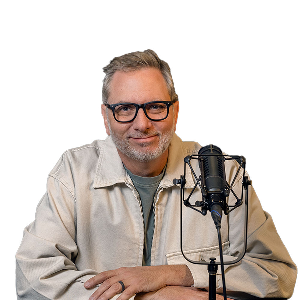 Rusty George smiling behind a microphone wearing a beige jacket.