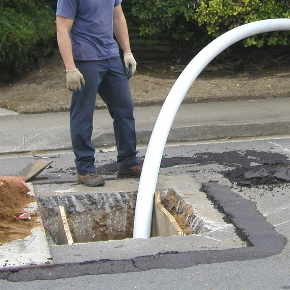 Worker stands next to open road hole with large white pipe installed for drain cleaning.