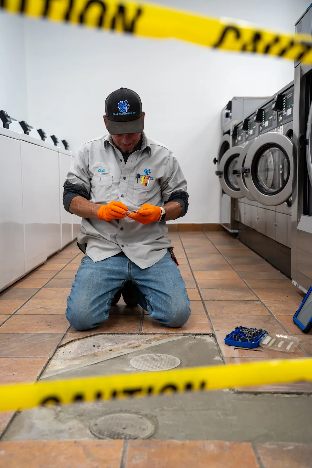 Plumber kneeling on tiled floor in laundry area, working near open floor drain with caution tape.